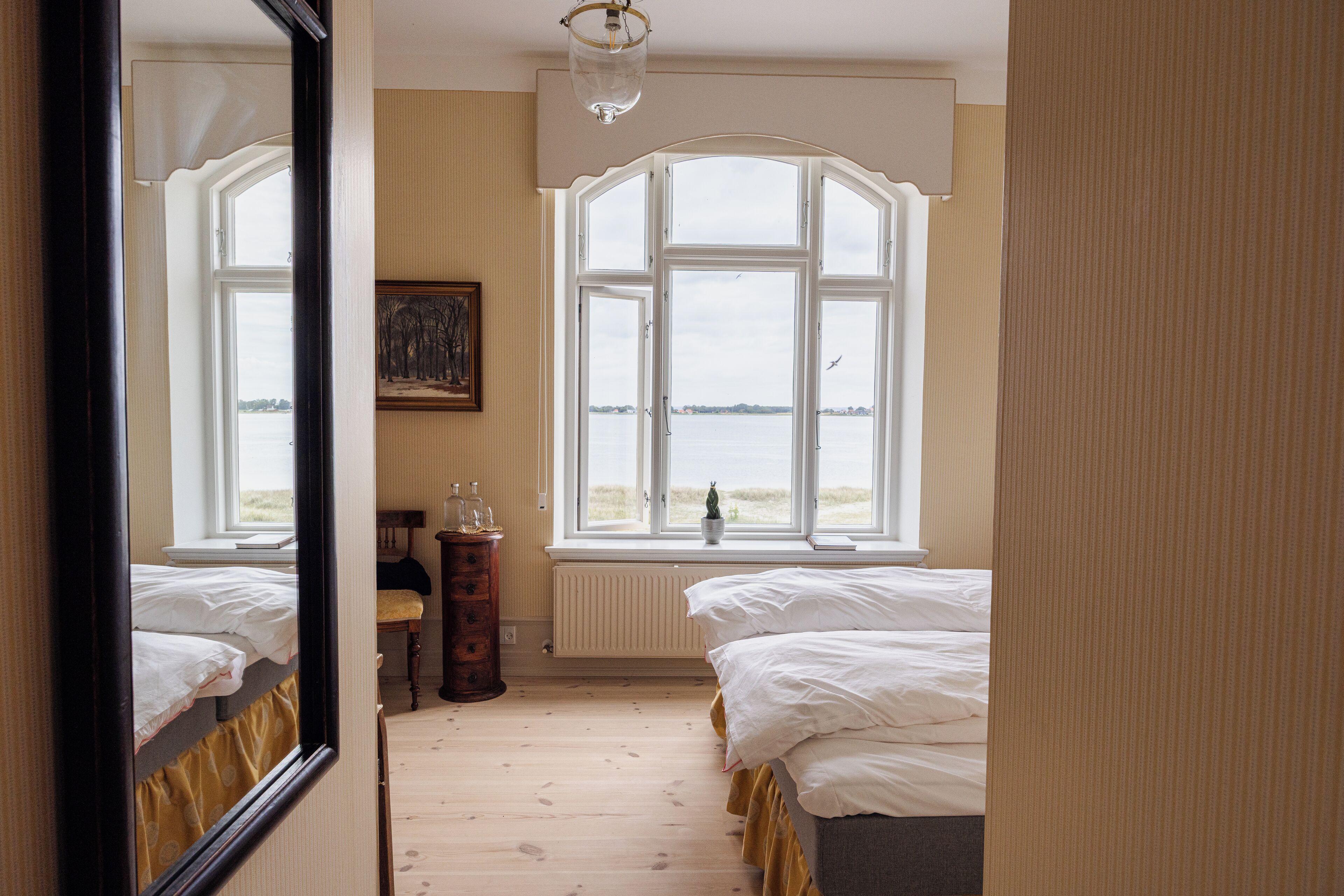 A bedroom with two beds, a large window with a view of the sea, a radiator, and a mirror reflecting the room.