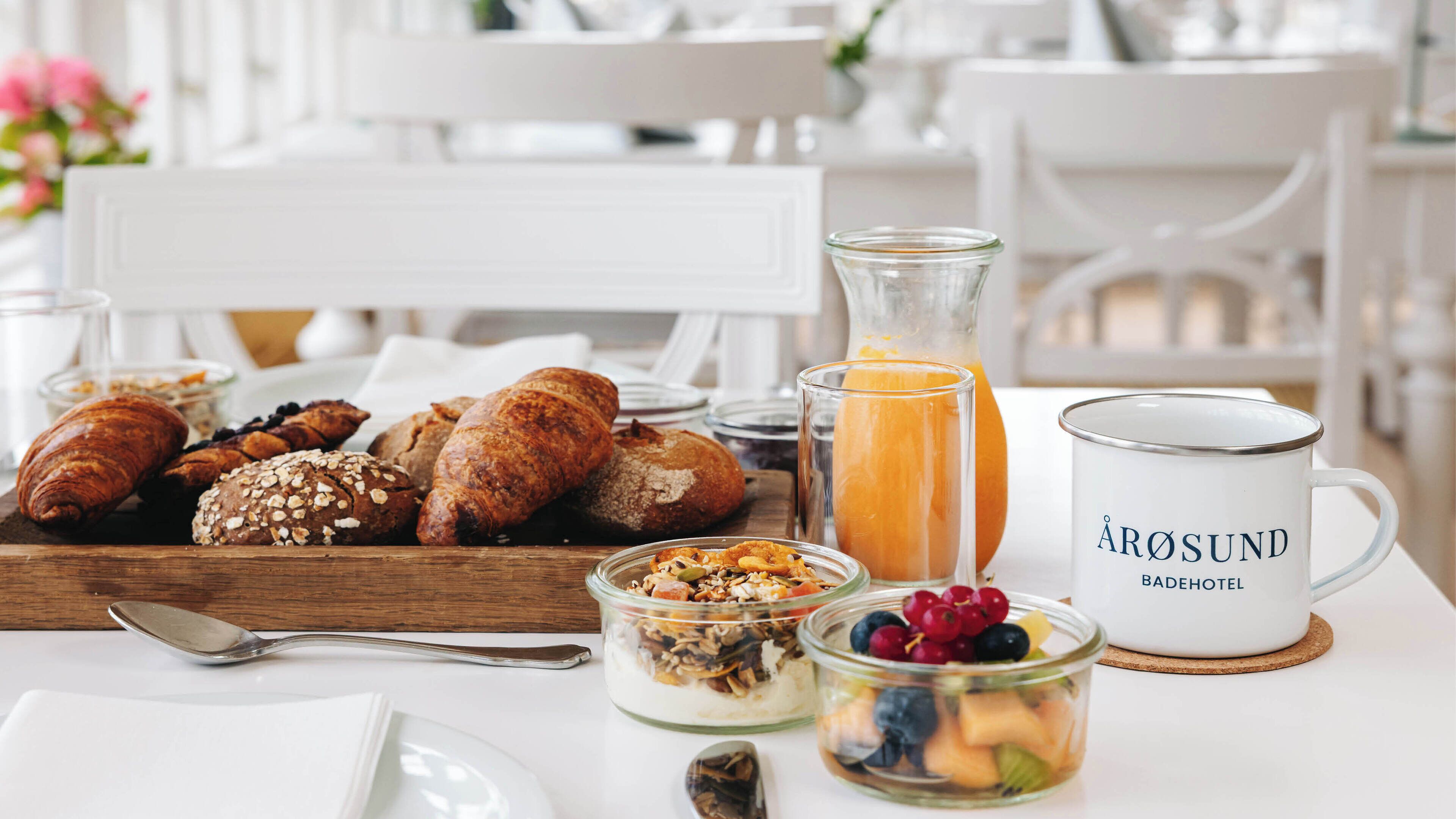 Desayuno con bollos, un vaso de zumo de naranja, una taza y un bol con fruta y frutos secos.