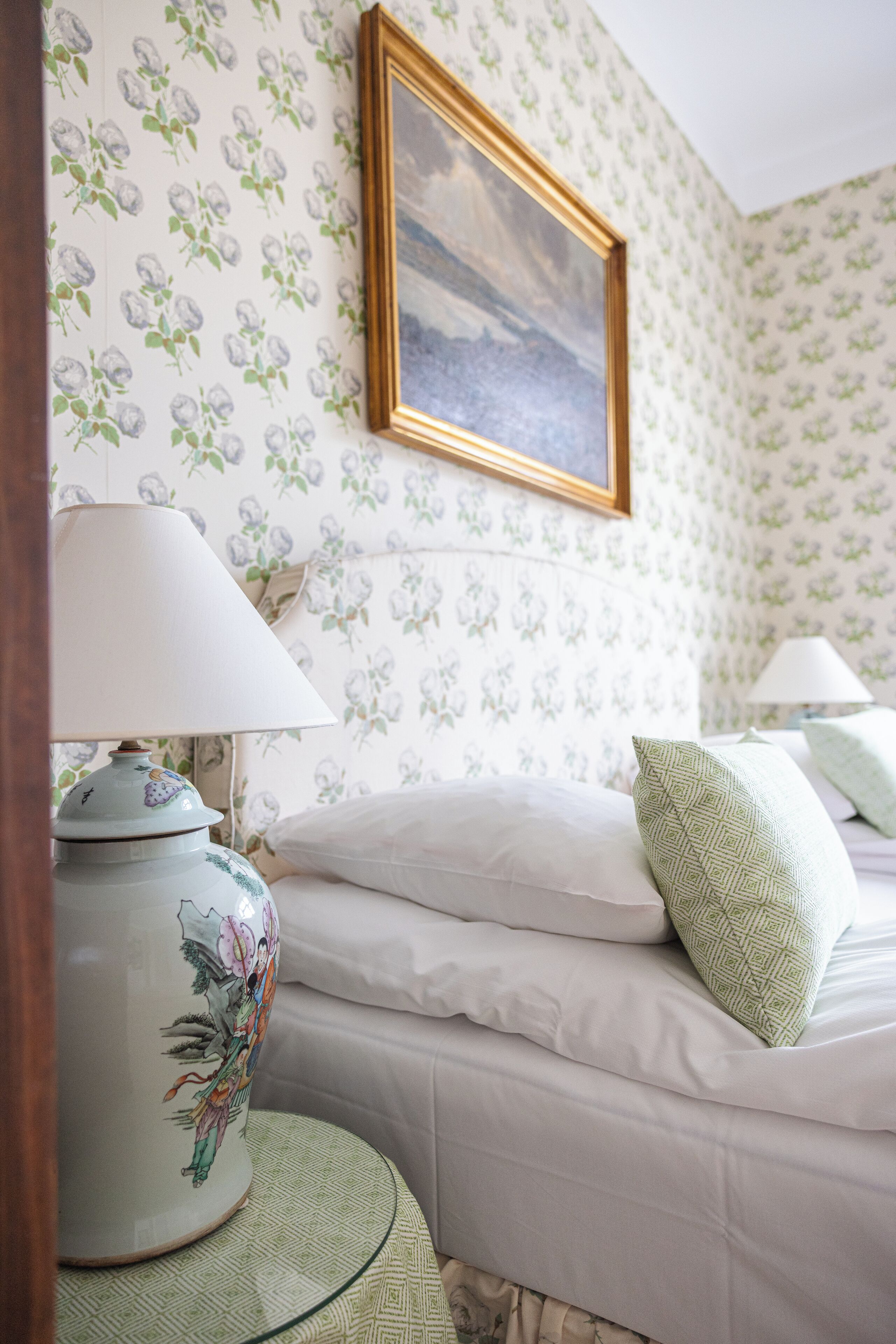 A bedroom with floral wallpaper, a bed with white bedding, a bedside table with a lamp, and a framed picture on the wall.