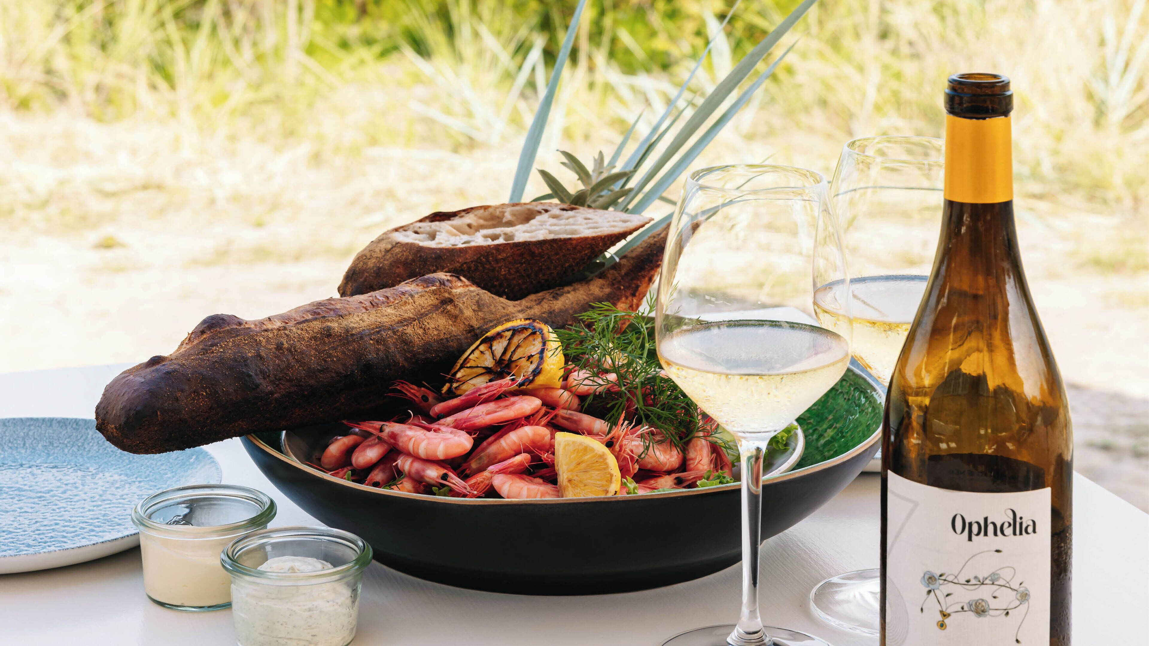 A seafood platter with shrimp, a grilled lemon, and a coconut on a black plate, accompanied by a bottle of wine and glasses.