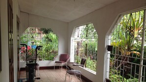 A sunlit room with a tiled floor, a white chair, a table, and a view of lush greenery through a barred window.