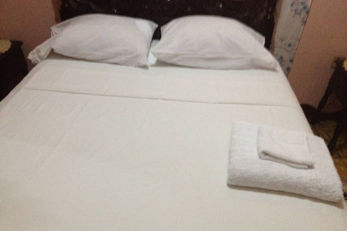 A neatly made bed with white linens and pillows, a folded white towel, and a dark headboard.