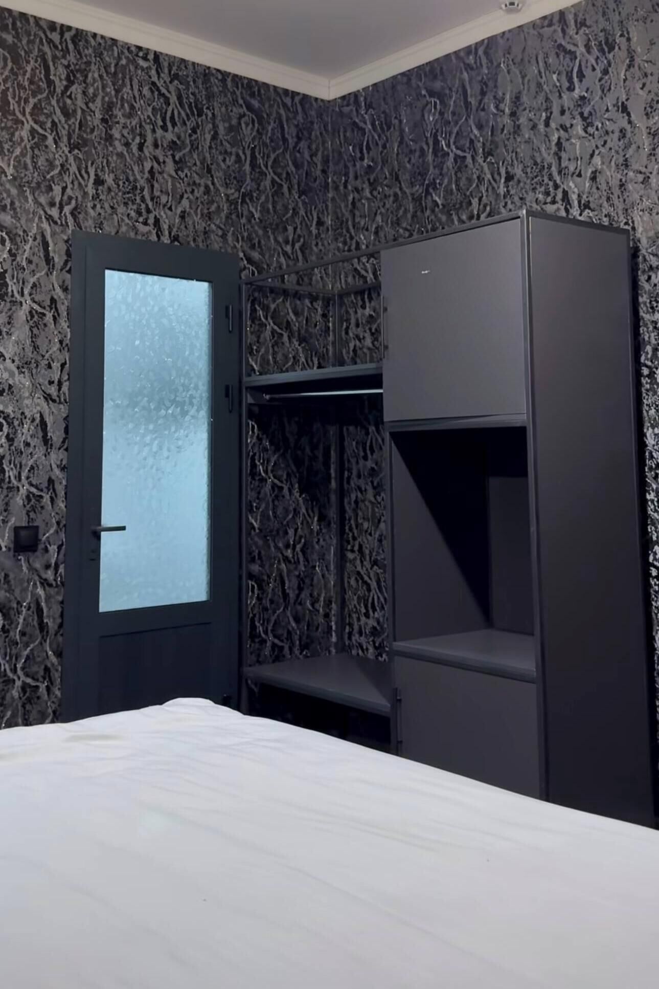 A compact room with a bed, a wardrobe, and a built-in bench with a glass door.