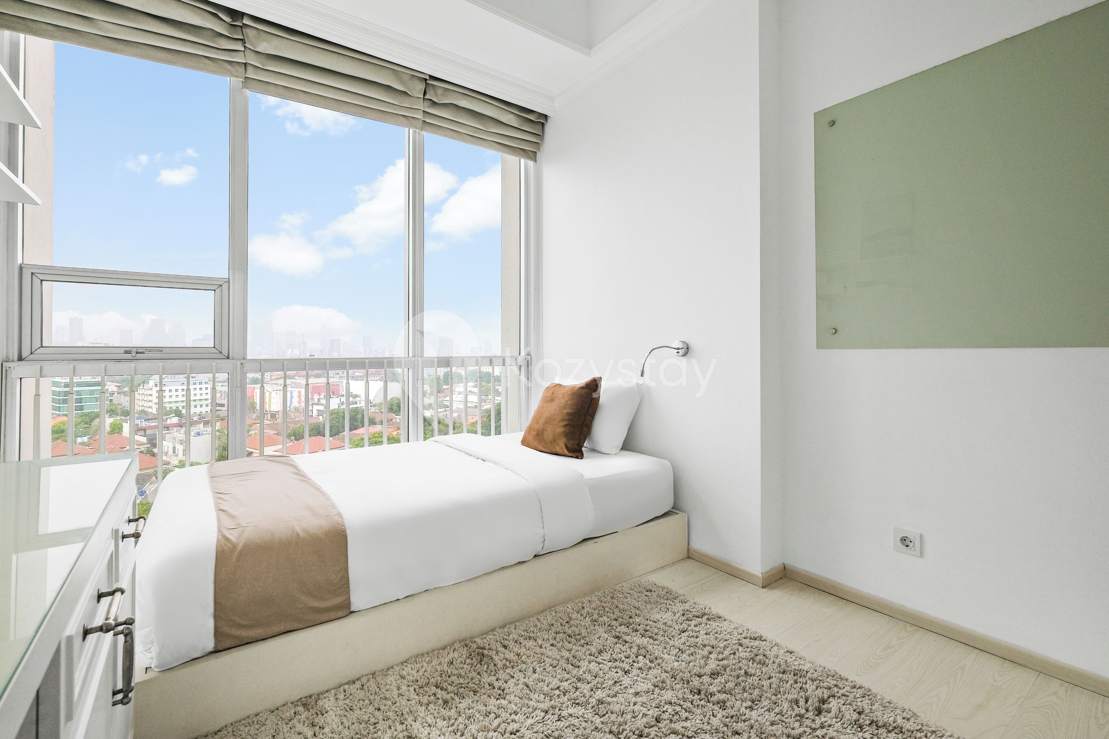 Un dormitorio moderno con un ventanal grande, una cama con ropa de cama blanca y una almohada marrón, una pared verde y vistas de edificios al exterior.