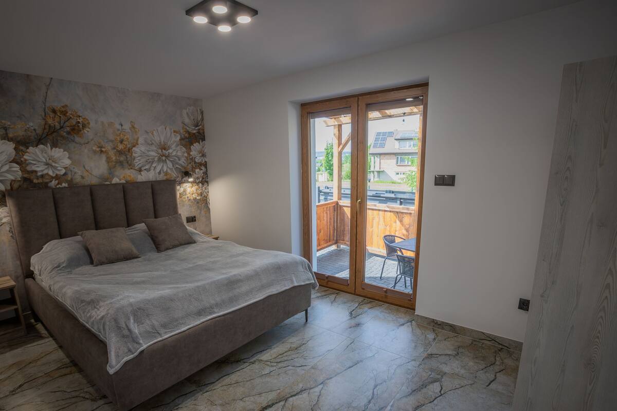 Een slaapkamer met een groot bed, een houten hoofdbord, een bloemen behang en een glazen schuifdeur naar een buitenruimte met een tafel en stoelen.