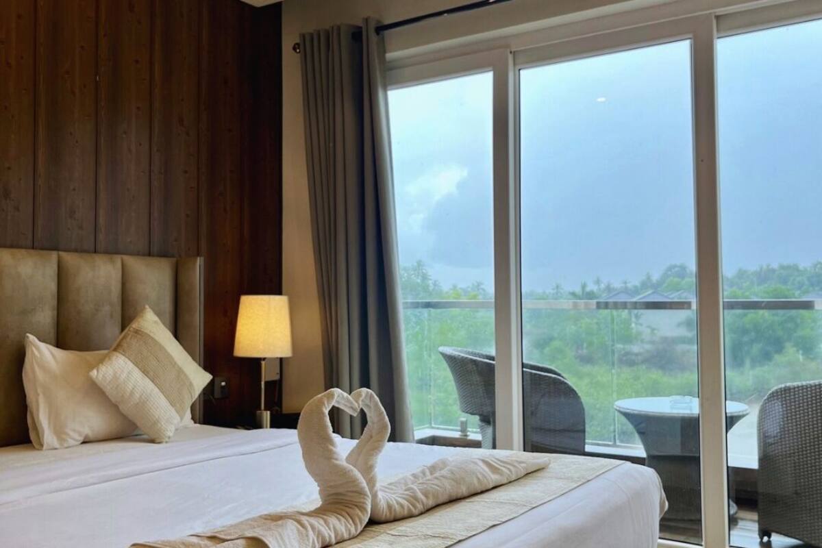 A hotel room with a large bed, a nightstand with a lamp, and a view of the outdoors through a large window.