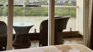 A hotel room with a large sliding glass door leading to a balcony overlooking a body of water and a hilly landscape. There is a bed with a white bedspread and a decorative pillow. Two wicker chairs are placed on the balcony. - PAS RESORTS (Anjuna)