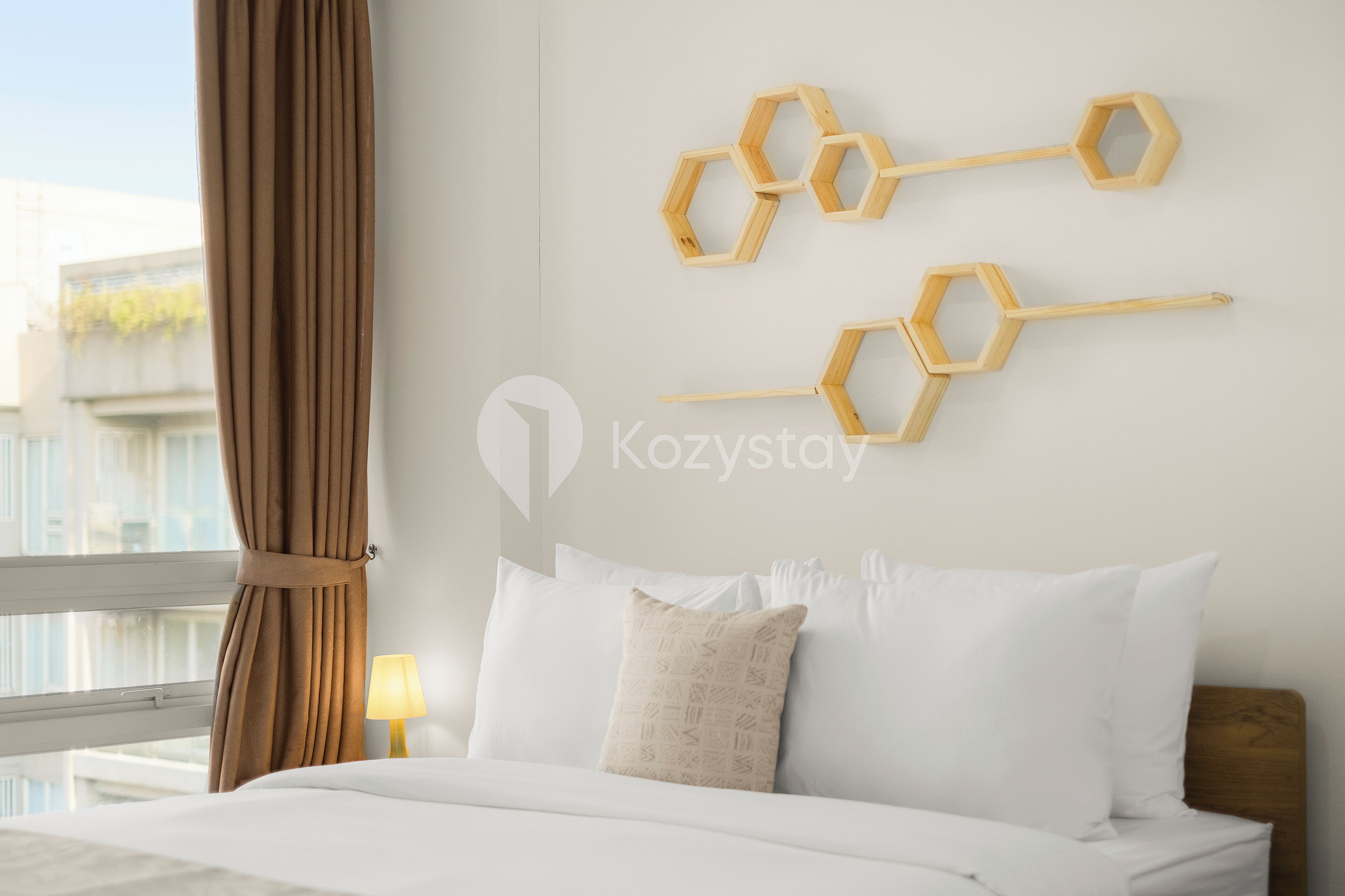 A hotel room with a bed, white bedding, a wooden headboard, and a wall-mounted hexagonal wooden shelf.