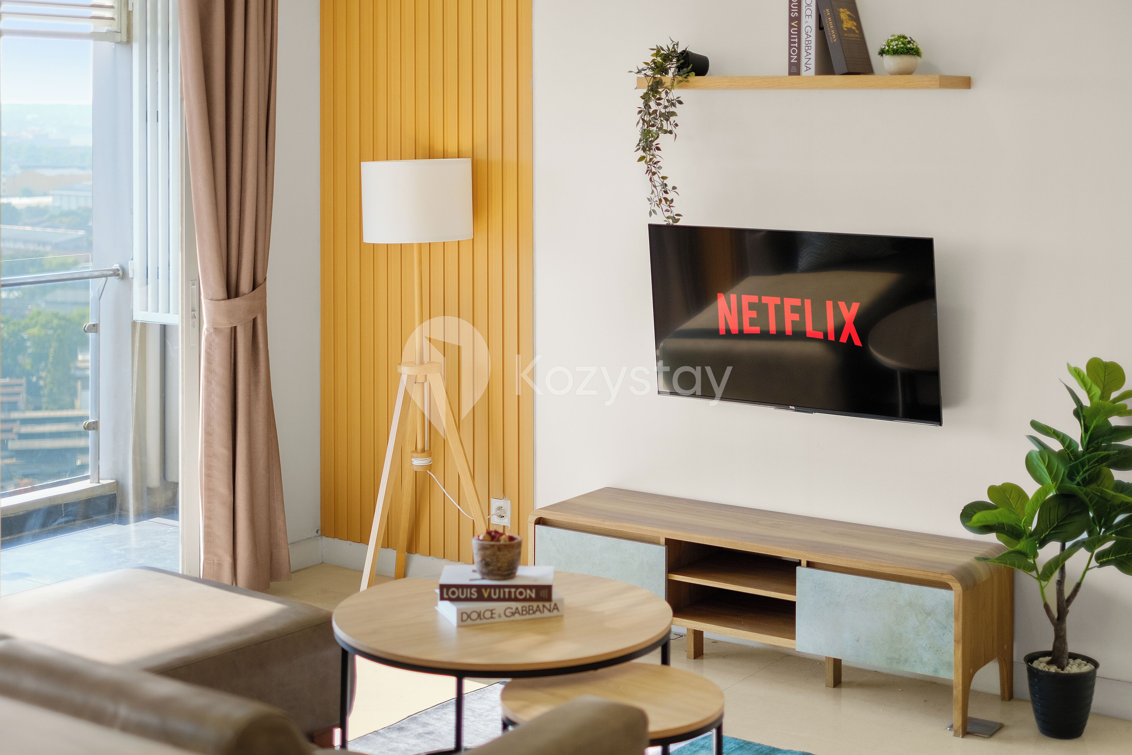 A modern living room with a television displaying "NETFLIX," a wooden bench, a small table with a book, and a potted plant.