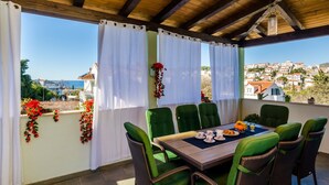 Un área de comedor al aire libre techada con sillas verdes y una mesa preparada para dos, con vista a un pueblo costero.