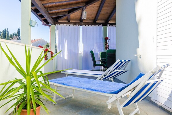 Ein überdachter Außenbereich mit zwei Liegestühlen und einem Tisch, einer Topfpflanze und Blick auf ein Haus im Hintergrund.