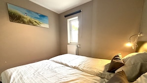 Een slaapkamer met twee bedden, een raam, een radiator en een schilderij aan de muur.