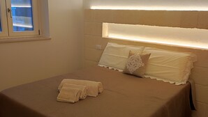 A neatly made bed with a headboard, a window with blinds, and a bedside table with a lamp.