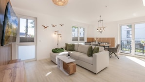 Un vaste espace de vie comprenant un canapé, une table basse et une table à manger. De grandes fenêtres, un balcon et un lustre moderne complètent l’ensemble.