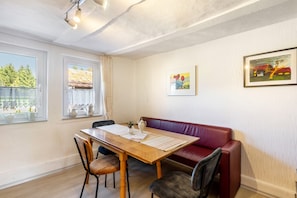 A dining area with a wooden table, chairs, a red leather bench, and two windows with curtains.