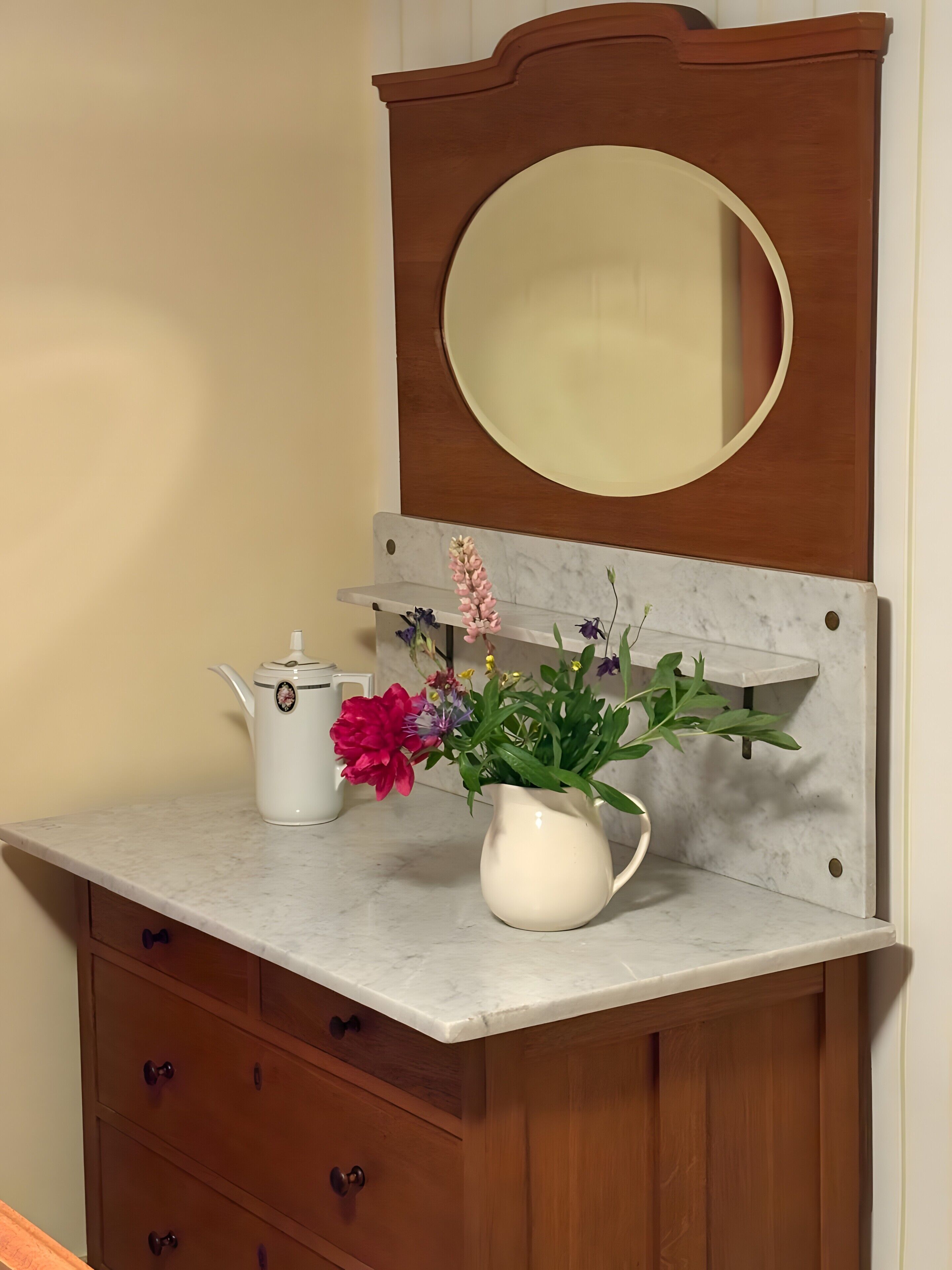 Un meuble-lavabo de salle de bain avec un miroir, un pichet blanc et un arrangement floral.