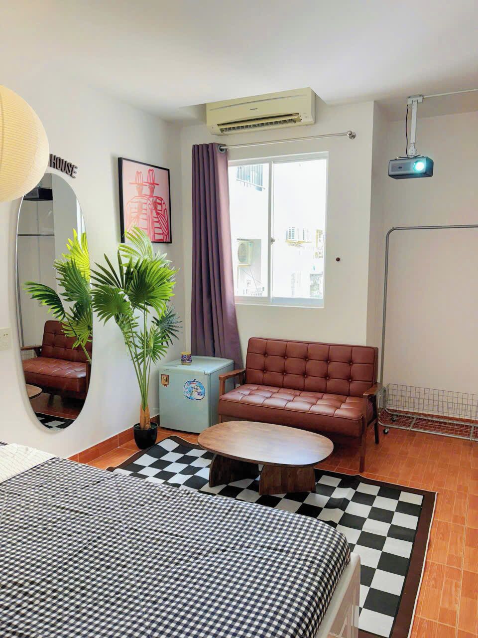 Habitación de hotel con piso a cuadros, un sofá, una mesita, una planta en maceta, un espejo y una ventana con cortinas.