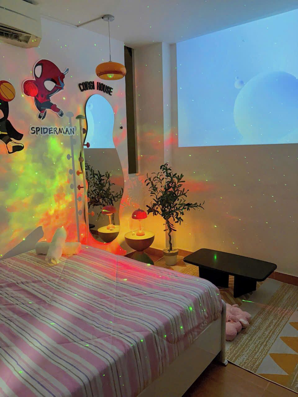 A bedroom with a bed, a projector screen, a wall mural, a small table, and a potted plant.