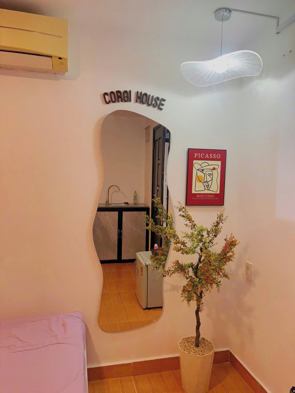 Una habitación con una cama, una planta en maceta, un aire acondicionado de pared, un accesorio de luz en el techo, un letrero que dice 'CORGI HOUSE' y una obra de arte de Picasso enmarcada.