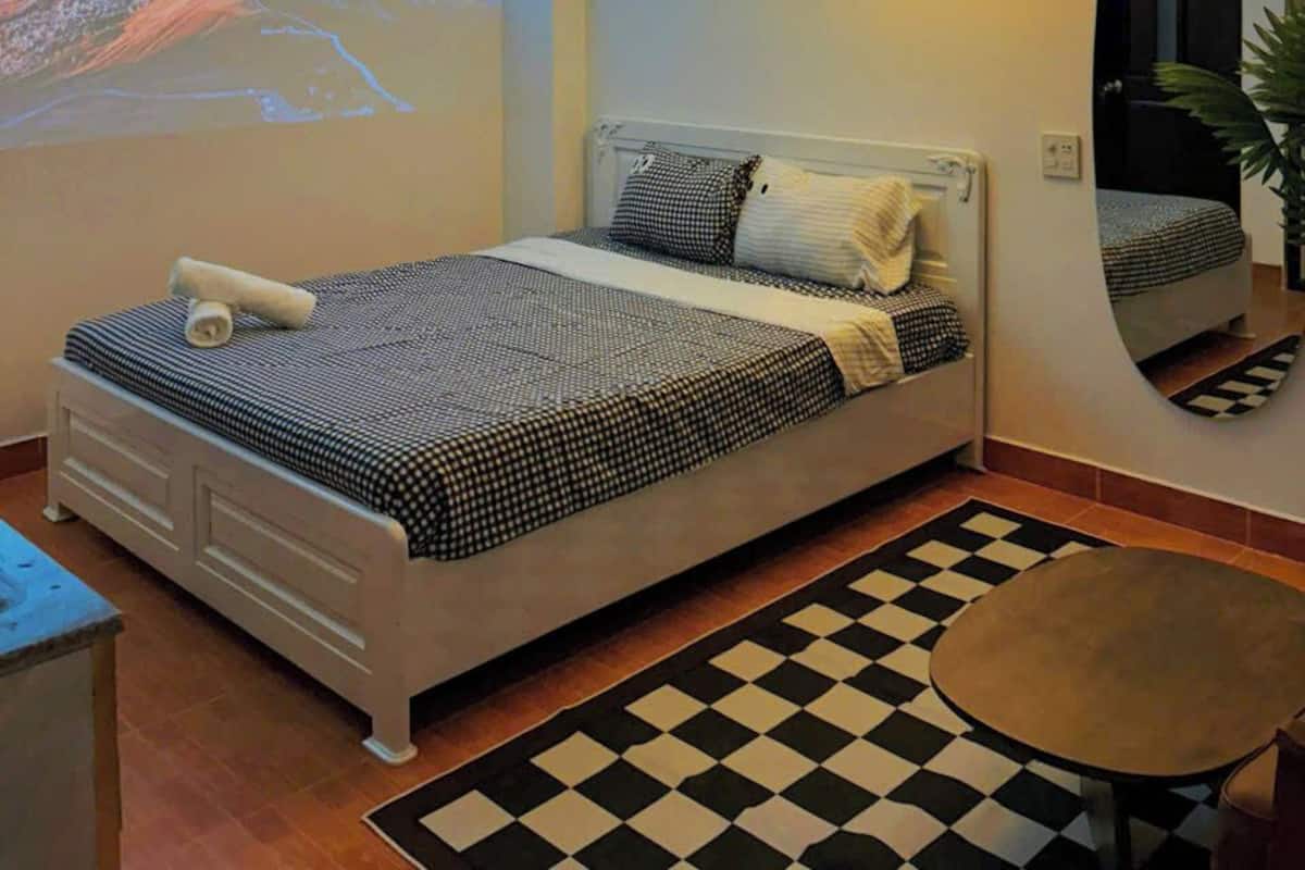 A bedroom with a bed, a checkered rug, a round table, a wall-mounted lamp, a projection of mountains, and a mirror with a door.