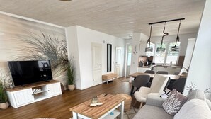 A modern living room with a large sofa, a coffee table, and a television. There is a dining area with a table and chairs visible in the background.