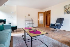 A living room with a sofa, two tables with pink mats, a chair, a bookshelf, and a door leading to another room.