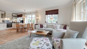 A spacious living area with a sofa, armchair, and dining table. There is a kitchen in the background with cabinets and a window. - Sölring Juwel Whg. 1 - Relaxed living in the Sölring Juwel Whg. 1 in (Wenningstedt-Braderup (Sylt))