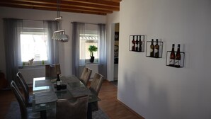 Un coin repas avec une table en verre, des chaises, un luminaire suspendu, une plante en pot et des bouteilles de vin exposées sur le mur.