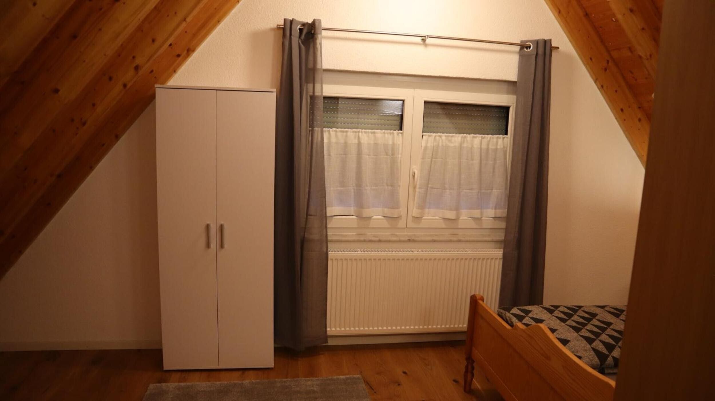 A bedroom with a sloped ceiling, a wardrobe, a radiator, a window with curtains, and a bed with a patterned blanket.