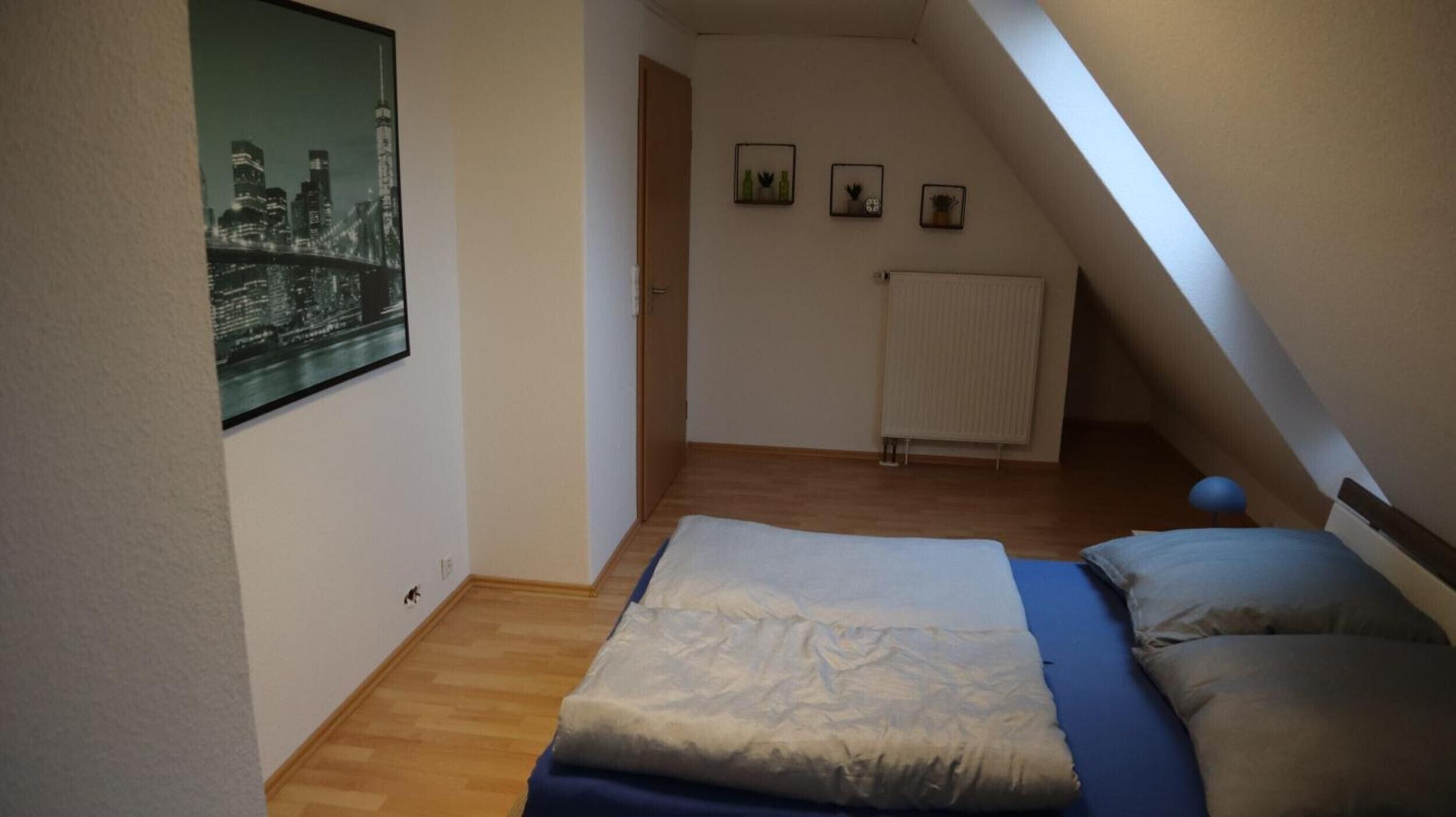 Ein kleines, ordentliches Schlafzimmer mit einem Einzelbett, einem Heizkörper und einem Stadtbild-Poster an der Wand.