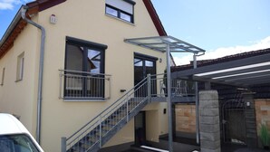 Un bâtiment à deux étages doté d’un espace extérieur abrité, d’un escalier et d’une voiture garée devant.