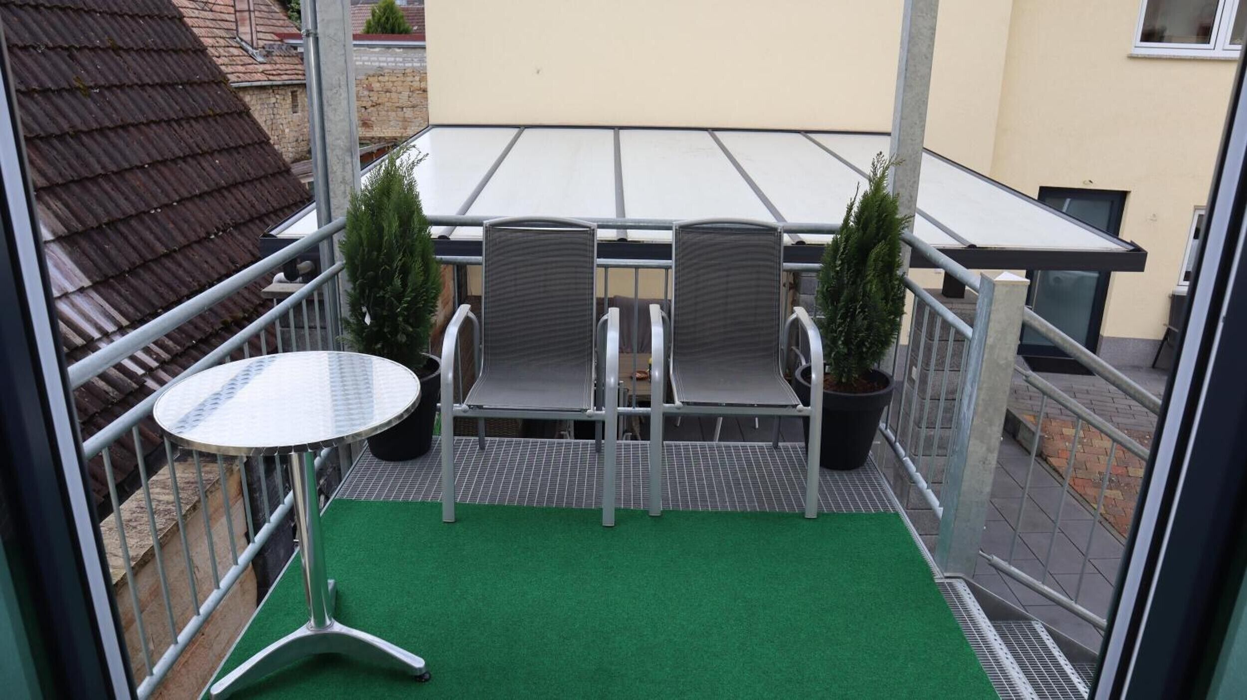 Un balcon avec une table et deux chaises, des plantes en pot et un sol recouvert d’un gazon artificiel vert.