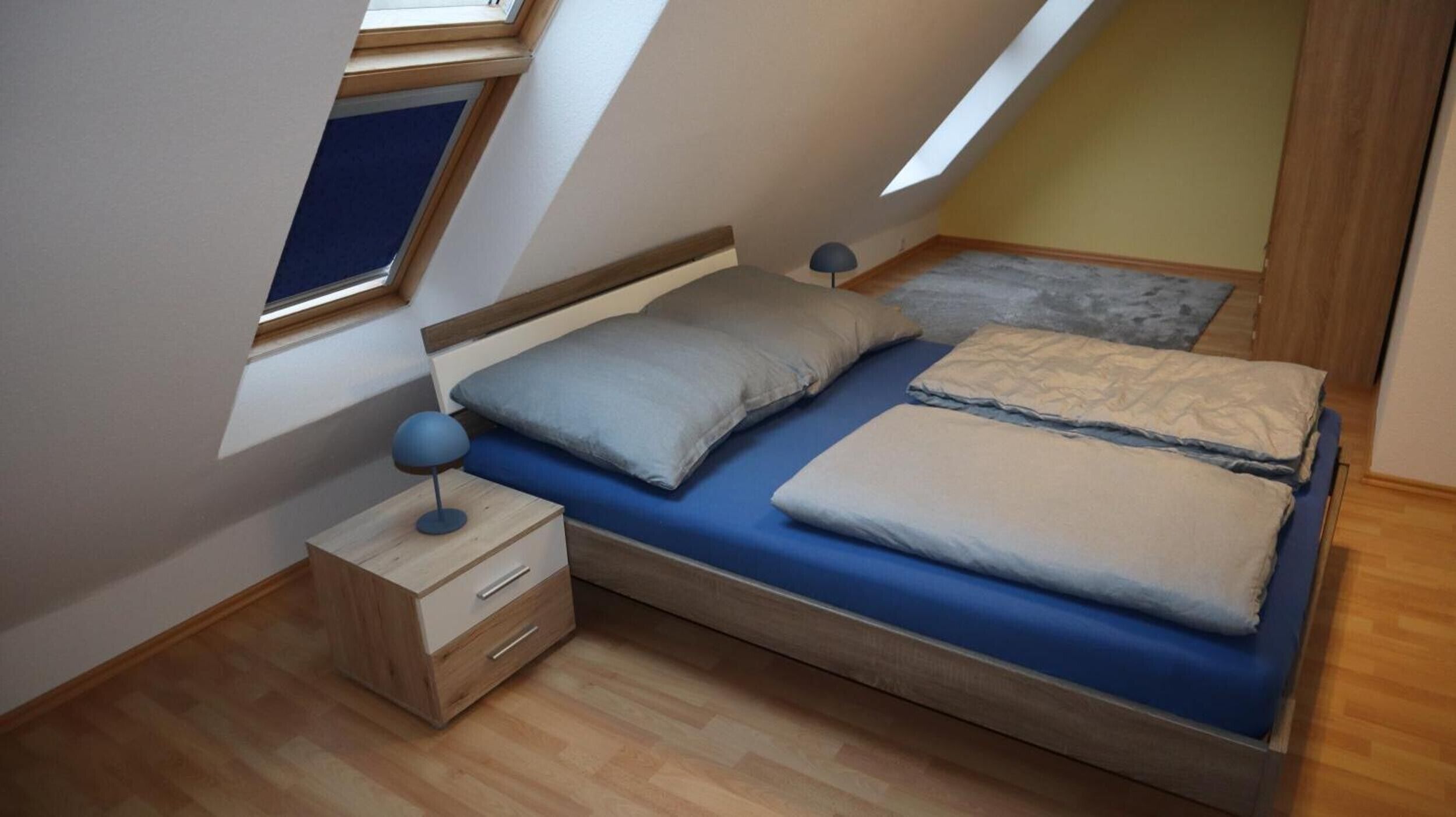 Une chambre avec un plafond en pente, un lit avec une literie bleue, une table de chevet avec une lampe et un petit tiroir.