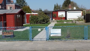 Ein durch Zäune umgebenes Grundstück mit einem roten Gebäude, einer weißen Garage und einem Schild mit der Aufschrift 'Ferienbungalows' und Kontaktinformationen.
