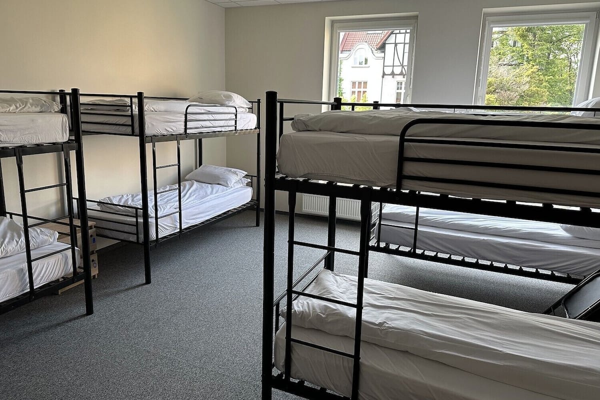 Une chambre de dortoir avec des lits superposés, un radiateur et des fenêtres donnant sur un bâtiment et des arbres.