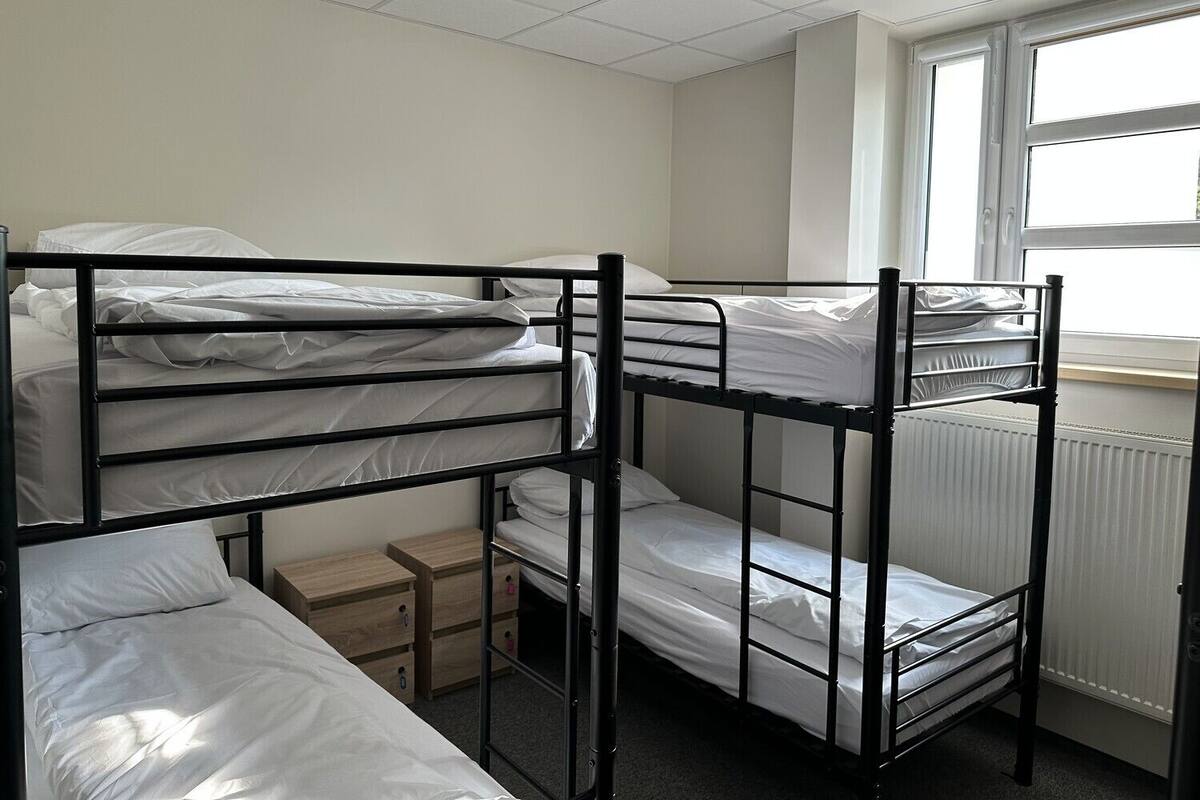 Une chambre de dortoir avec des lits superposés, une petite commode en bois et une fenêtre avec des stores.
