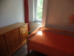 Une chambre avec une commode en bois, un lit simple, une fenêtre donnant sur un paysage verdoyant et un radiateur.