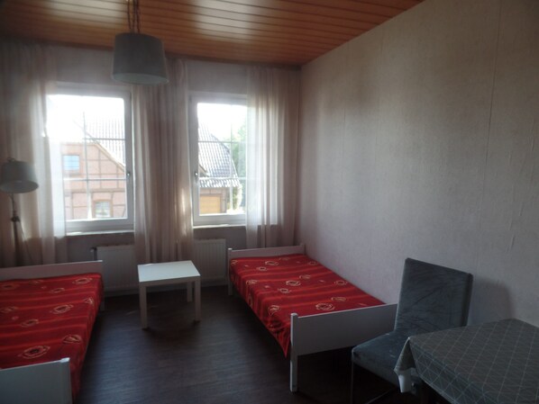 A small, simple room with two single beds, a small table, and a chair.