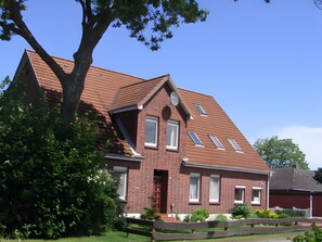 A red brick house with a sloped roof, multiple windows, and a large tree in the foreground. - Apartment '3-Raum' with Shared Terrace and Wi-Fi (Cuxhaven)