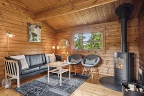 A log cabin interior with a sofa, chairs, a table, a fireplace, and a window with a view of greenery.