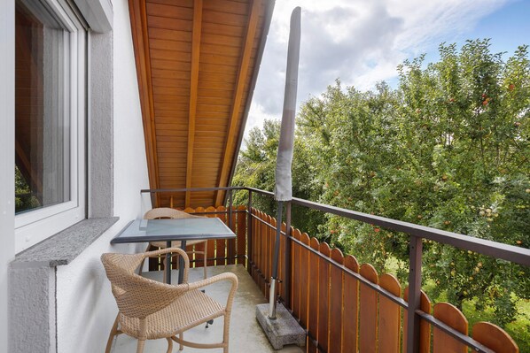 Un balcón con mesa y sillas, barandilla de madera y vista a árboles.
