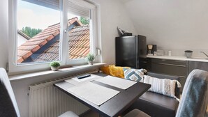 Een kleine kamer met een bureau, een stoel, een bank, een radiator, een raam met zonwering, een klein plantje en een televisie.