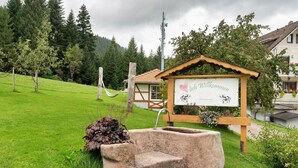 Une enseigne rustique en bois, ornée d’un cœur et d’un chien, un puits en pierre et un hamac, le tout installé dans un espace verdoyant avec des arbres et une maison en arrière-plan.
