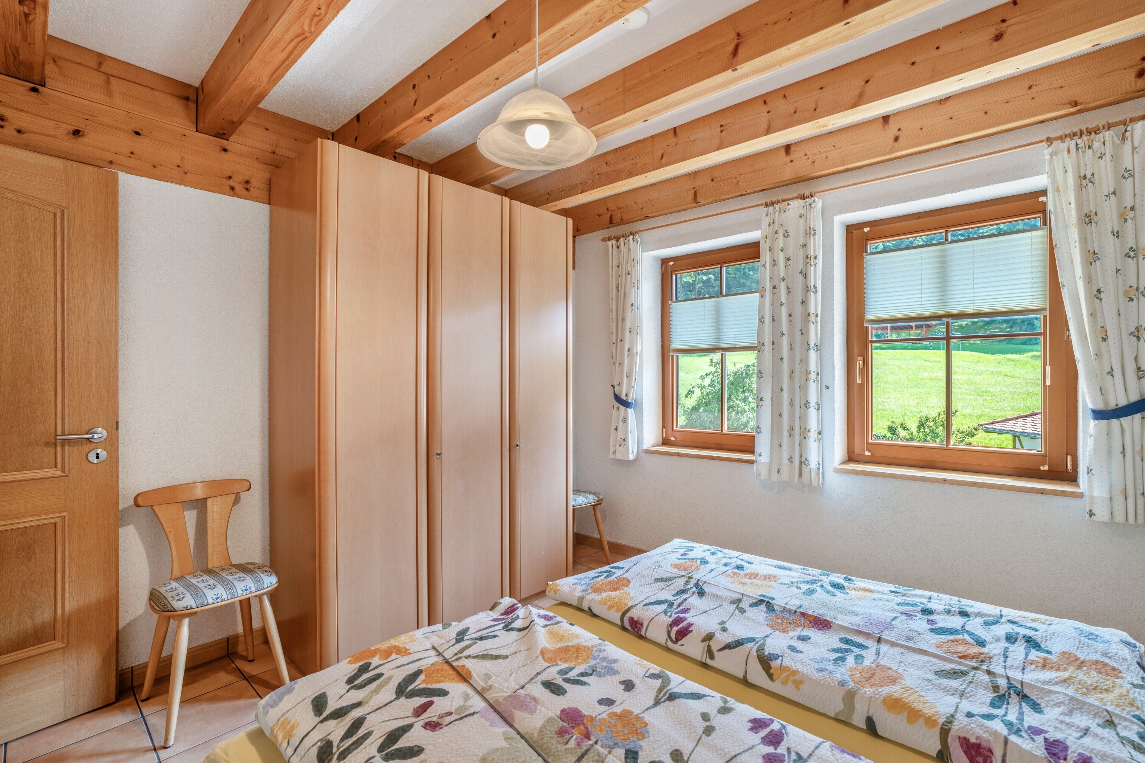 A room with wooden beams, a bed with floral bedding, a chair, a wardrobe, and a window with curtains.
