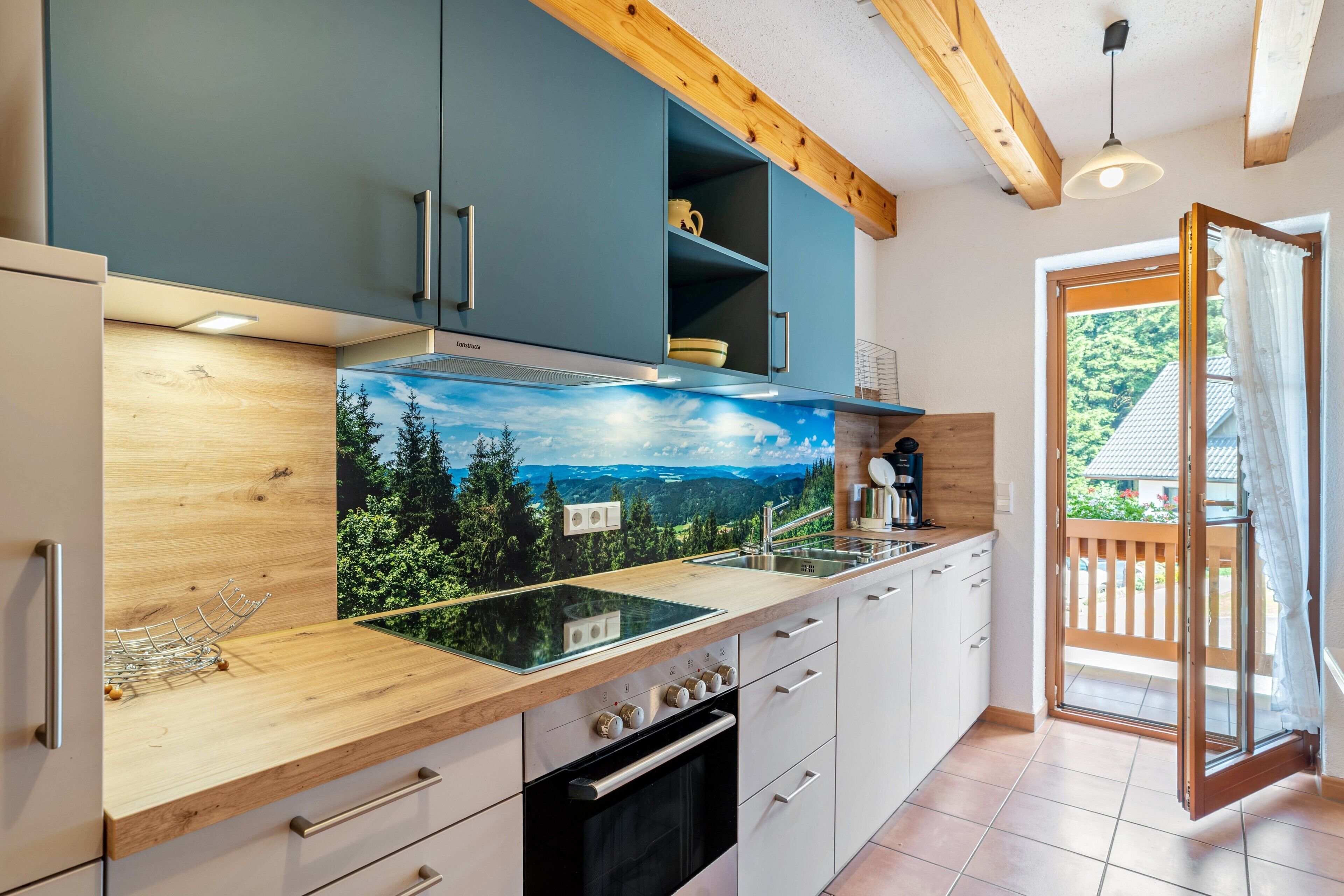 Une cuisine moderne avec des meubles en bois, une cuisinière encastrée et une vue sur l’extérieur grâce à une porte vitrée.