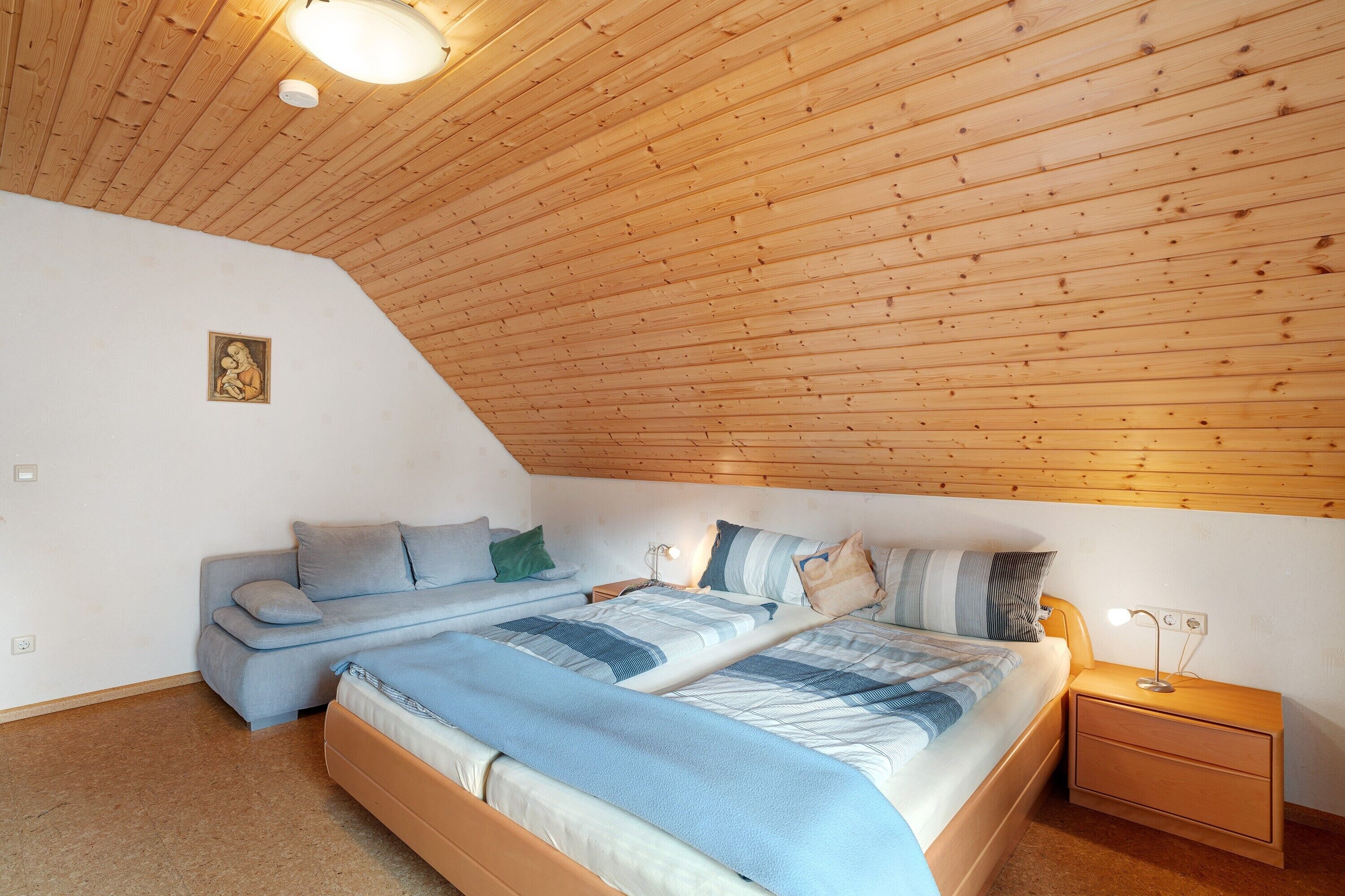 A bedroom with a sloped ceiling, a bed with a blue and white striped bedspread, a sofa with cushions, a bedside table with a lamp, and a small framed picture on the wall.
