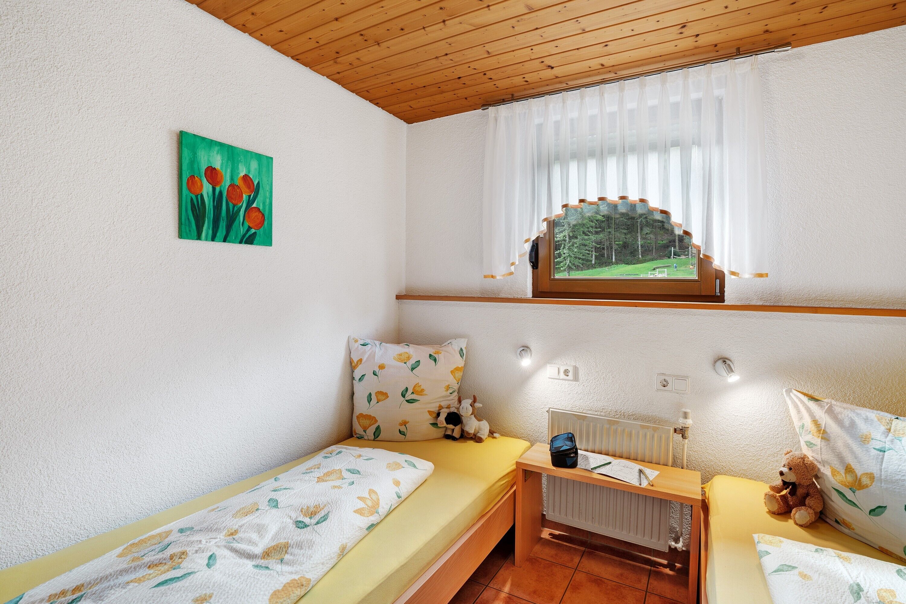 Ein kleiner, gemütlicher Raum mit zwei Einzelbetten, einem Fenster mit Blick auf eine grüne Landschaft, einem Heizkörper und einem kleinen Tisch mit einem Teddybär.