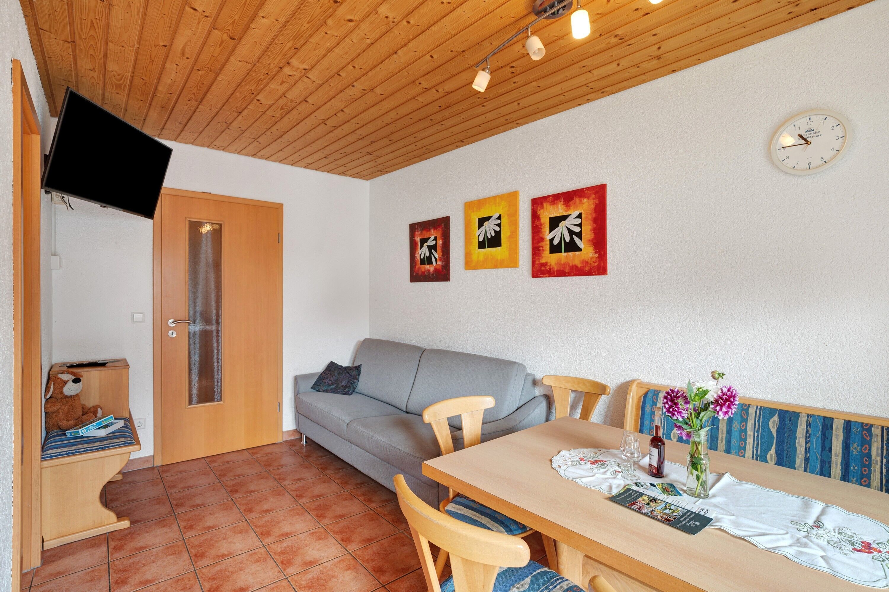 A room with a wooden ceiling, a gray sofa, a dining table with chairs, a wall clock, and three framed artworks.