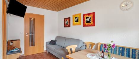 A room with a wooden ceiling, a gray sofa, a dining table with chairs, a wall clock, and three framed artworks.