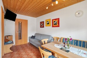 A room with a wooden ceiling, a gray sofa, a dining table with chairs, a wall clock, and three framed artworks.
